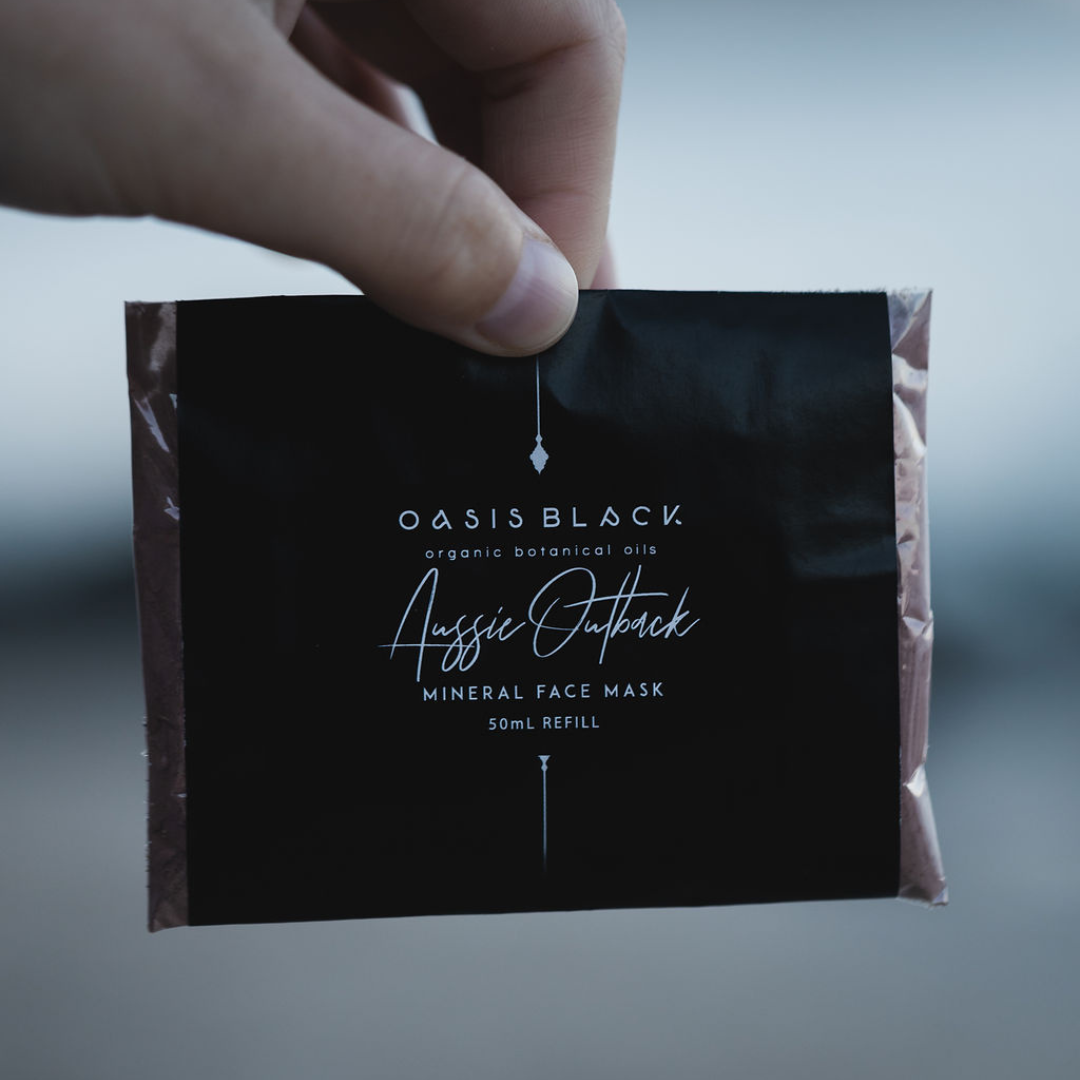 Hand holding a plastic free & home compostable sachet of Oasis Black's Powdered Clay Mud Mask in Aussie Outback - a red dirt coloured facial mask activated with water featuring Australian Clays, Davidson Plum, Lemon Myrtle, Muntrie, Kakadu Plum & Black Seeds. Available exclusively from Oasis Black - makers of All Natural and Organic Botanical Skincare featuring Black Seeds (Nigella Sativa). All products are handmade in small batches to waterless formulations in the Byron Bay hinterland