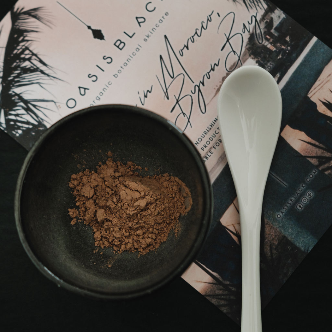 Black clay cosmic mixing bowl & teaspoon with terracotta coloured powder sitting on Oasis Black branded flyer. The powder is Oasis Black's Mineral Mud Mask in Mediterranean that's activated with water & featuring Mediterranean Clays, Rose Petals, Hibiscus, Aloe Vera, Calendula & Black Seeds. Available exclusively from Oasis Black - makers of All Natural and Organic Botanical Skincare featuring Black Seeds (Nigella Sativa). Handmade in small batches to waterless formulations in the Byron Bay Hinterland. 