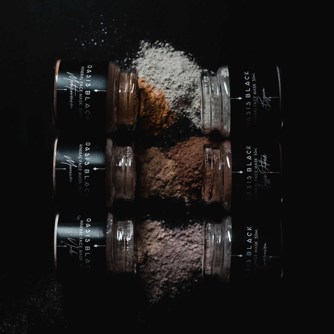 6 x 50ml glass jars labelled with Oasis Black branding, lying on their sides with mineral clay Mud Mask powder in different colours spilling out of each jar. Oasis Black are makers of All Natural and Organic Botanical Skincare featuring Black Seeds (Nigella Sativa). Handmade in small batches to waterless formulations in the Byron Bay Hinterland. 