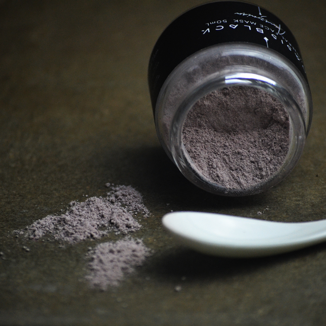 Glass jar of Oasis Black's Amazonian Mineral Clay Mud Mask lying on it's side with a teaspoon and the powder spilled out on a grey surface. Amazonian is a purple coloured mask activated with water featuring Amazonian Clays, Acai, Maca, Inca berries, Camu Camu and Black Seeds. Available exclusively from Oasis Black - makers of All Natural and Organic Botanical Skincare featuring Black Seeds (Nigella Sativa). All products are handmade in small batches to waterless formulations in the Byron Bay hinterland. 