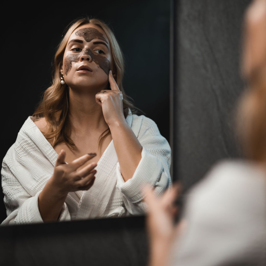 Woman in a white bathrobe looking in a mirror applying Oasis Black's Amazonian Mineral Clay Mud Mask with her fingers. Amazonian is a purple coloured facial mask activated with water featuring Amazonian Clays, Acai, Maca, Inca berries, Camu Camu and Black Seeds. Available exclusively from Oasis Black - makers of All Natural and Organic Botanical Skincare featuring Black Seeds (Nigella Sativa). All products are handmade in small batches to waterless formulations in the Byron Bay hinterland