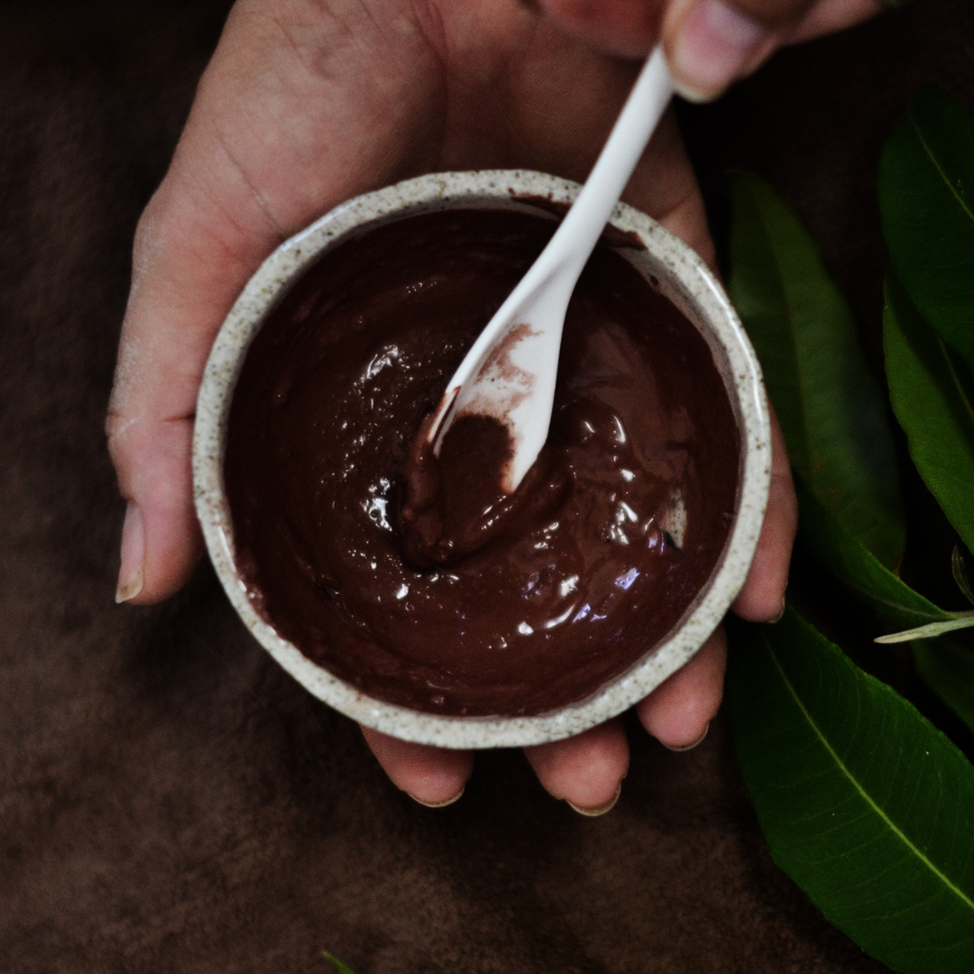 Hand holding a small mixing bowl of red clay paste with a spoon, surrounded by green leaves. The paste is Oasis Black's Aussie Outback Mineral Mud mask that's activated with water and features Australian Clays, Davidson Plum, Lemon Myrtle, Muntrie, Kakadu Plum & Black Seeds. Available exclusively from Oasis Black - makers of All Natural and Organic Botanical Skincare featuring Black Seeds (Nigella Sativa). All products are handmade in small batches to waterless formulations in the Byron Bay hinterland