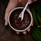 Hand holding a small mixing bowl of red clay paste with a spoon, surrounded by green leaves. The paste is Oasis Black's Aussie Outback Mineral Mud mask that's activated with water and features Australian Clays, Davidson Plum, Lemon Myrtle, Muntrie, Kakadu Plum & Black Seeds. Available exclusively from Oasis Black - makers of All Natural and Organic Botanical Skincare featuring Black Seeds (Nigella Sativa). All products are handmade in small batches to waterless formulations in the Byron Bay hinterland