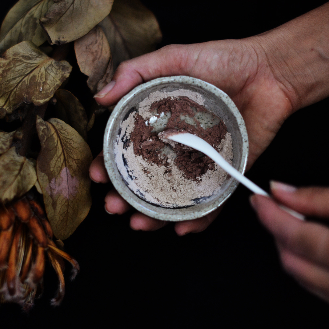 Person holding a mixing bowl of clay powder with a spoon, surrounded by green leaves. The powder is Oasis Black's Aussie Outback Mineral Mud mask that's being activated with water and features Australian Clays, Davidson Plum, Lemon Myrtle, Muntrie, Kakadu Plum & Black Seeds. Available exclusively from Oasis Black - makers of All Natural and Organic Botanical Skincare featuring Black Seeds (Nigella Sativa). All products are handmade in small batches to waterless formulations in the Byron Bay hinterland