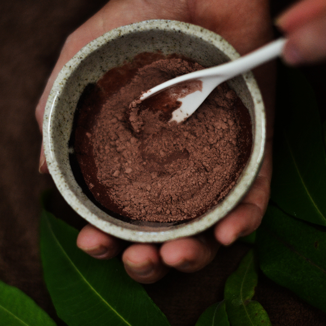 Person holding a mixing bowl of clay powder with a spoon, surrounded by green leaves. The powder is Oasis Black's Aussie Outback Mineral Mud mask that's being activated with water and features Australian Clays, Davidson Plum, Lemon Myrtle, Muntrie, Kakadu Plum & Black Seeds. Available exclusively from Oasis Black - makers of All Natural and Organic Botanical Skincare featuring Black Seeds (Nigella Sativa). All products are handmade in small batches to waterless formulations in the Byron Bay hinterland