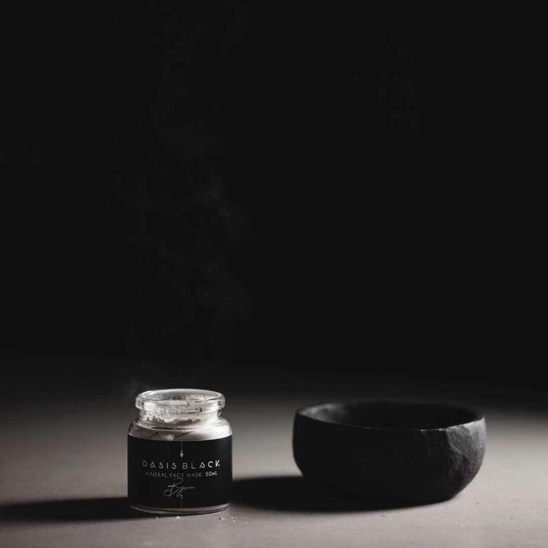 A jar of the Oasis Black Mineral Clay Mud Mask in Byron next to a black clay mixing bowl on a grey surface & black background. Byron is a stormy turquoise coloured mask featuring French Green Clay, Spirulina, Blue Butterfly Pea Flower, Lemon Myrtle and Black Seeds. Available exclusively from Oasis Black - makers of All Natural and Organic Botanical Skincare featuring Black Seeds (Nigella Sativa). Handmade in small batches to waterless formulations in the Byron Bay Hinterland. 
