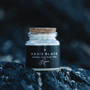 Glass jar (with a cork lid) of Oasis Black Mineral Face Mask in Byron on a dark rocky background. Byron is a stormy turquoise coloured mask featuring French Green Clay, Spirulina, Blue Butterfly Pea Flower, Lemon Myrtle and Black Seeds. Available exclusively from Oasis Black - makers of All Natural and Organic Botanical Skincare featuring Black Seeds (Nigella Sativa). Handmade in small batches to waterless formulations in the Byron Bay Hinterland. 