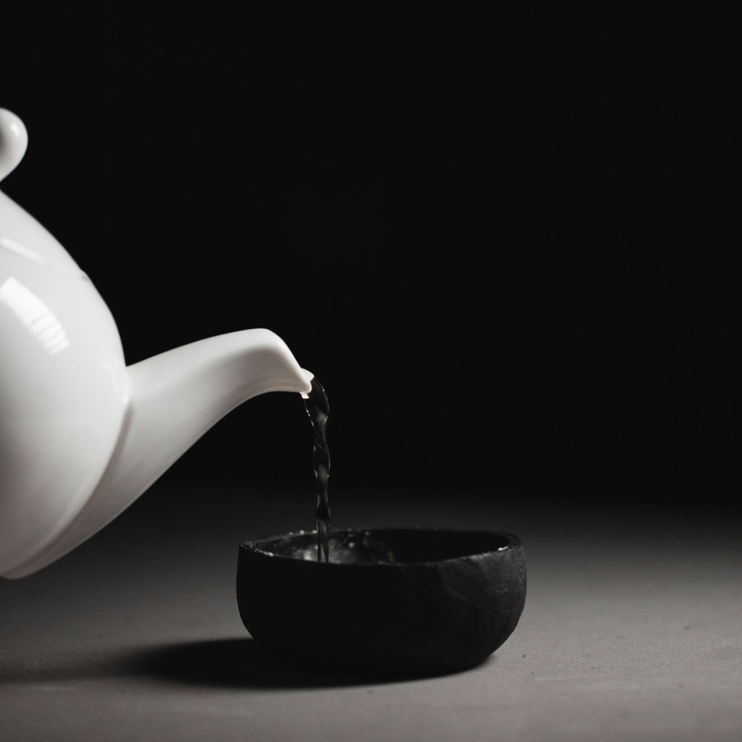 White teapot pouring water into a black clay mixing bowl containing Oasis Black's Mineral Clay Mud Mask in Byron, on a dark background. Byron is a stormy turquoise coloured mask featuring French Green Clay, Spirulina, Blue Butterfly Pea Flower, Lemon Myrtle and Black Seeds. Available exclusively from Oasis Black - makers of All Natural and Organic Botanical Skincare featuring Black Seeds (Nigella Sativa). Handmade in small batches to waterless formulations in the Byron Bay Hinterland.