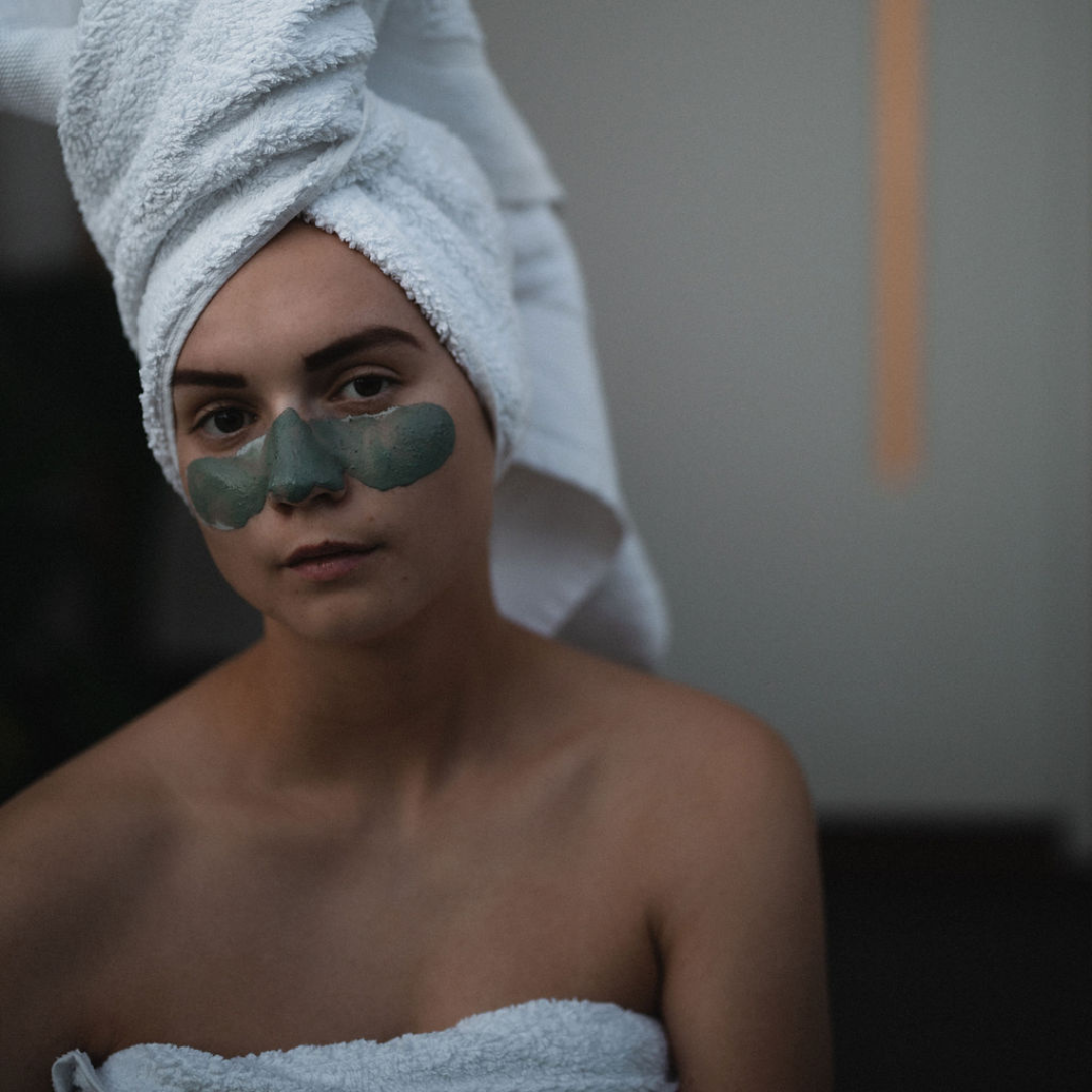 Woman with a green facial mask  applied to her nose and cheeks, wearing a towel on her head in a bathroom. The mask is Oasis Black's Mineral Clay Mud Mask in Byron - a stormy turquoise coloured mask featuring French Green Clay, Spirulina, Blue Butterfly Pea Flower, Lemon Myrtle and Black Seeds. Available exclusively from Oasis Black - makers of All Natural and Organic Botanical Skincare featuring Black Seeds (Nigella Sativa). Handmade in small batches to waterless formulations in the Byron Bay Hinterland.