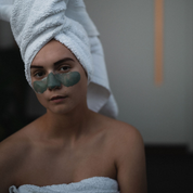 Woman with a green facial mask  applied to her nose and cheeks, wearing a towel on her head in a bathroom. The mask is Oasis Black's Mineral Clay Mud Mask in Byron - a stormy turquoise coloured mask featuring French Green Clay, Spirulina, Blue Butterfly Pea Flower, Lemon Myrtle and Black Seeds. Available exclusively from Oasis Black - makers of All Natural and Organic Botanical Skincare featuring Black Seeds (Nigella Sativa). Handmade in small batches to waterless formulations in the Byron Bay Hinterland.