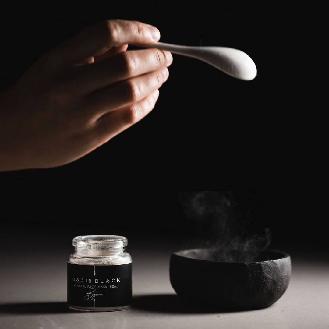 Hand holding a ceramic spoon over a black clay mixing bowl next to jar labelled Oasis Black Mineral Clay Mud Mask in Byron, with smoke rising from the bowl. Byron is a stormy turquoise coloured mask featuring French Green Clay, Spirulina, Blue Butterfly Pea Flower, Lemon Myrtle and Black Seeds. Available exclusively from Oasis Black - makers of All Natural and Organic Botanical Skincare featuring Black Seeds (Nigella Sativa). Handmade in small batches to waterless formulations in the Byron Bay Hinterland.