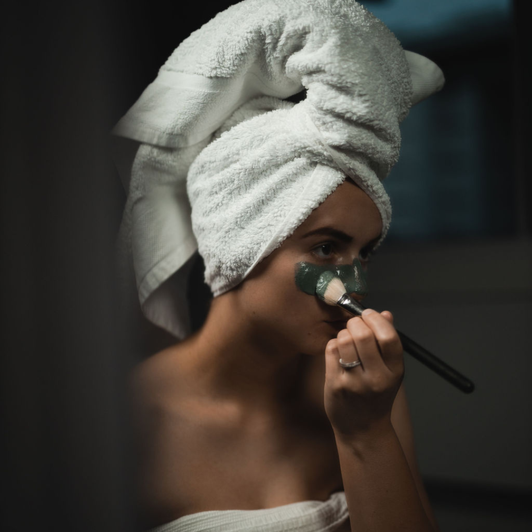 Woman applying a green facial mask with a brush, wearing a towel on her head. The mask is Oasis Black's Mineral Clay Mud Mask in Byron - a stormy turquoise coloured mask featuring French Green Clay, Spirulina, Blue Butterfly Pea Flower, Lemon Myrtle and Black Seeds. Available exclusively from Oasis Black - makers of All Natural and Organic Botanical Skincare featuring Black Seeds (Nigella Sativa). Handmade in small batches to waterless formulations in the Byron Bay Hinterland.