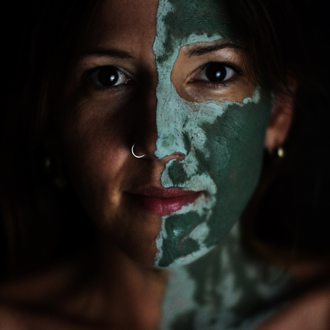 Woman with a green facial mask on one side of her face, with a dark background. The mask is Oasis Black's Mineral Clay Mud Mask in Byron - a stormy turquoise coloured mask featuring French Green Clay, Spirulina, Blue Butterfly Pea Flower, Lemon Myrtle and Black Seeds. Available exclusively from Oasis Black - makers of All Natural and Organic Botanical Skincare featuring Black Seeds (Nigella Sativa). Handmade in small batches to waterless formulations in the Byron Bay Hinterland.