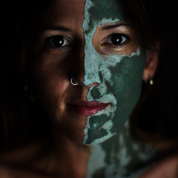 Woman with a green facial mask on one side of her face, with a dark background. The mask is Oasis Black's Mineral Clay Mud Mask in Byron - a stormy turquoise coloured mask featuring French Green Clay, Spirulina, Blue Butterfly Pea Flower, Lemon Myrtle and Black Seeds. Available exclusively from Oasis Black - makers of All Natural and Organic Botanical Skincare featuring Black Seeds (Nigella Sativa). Handmade in small batches to waterless formulations in the Byron Bay Hinterland.