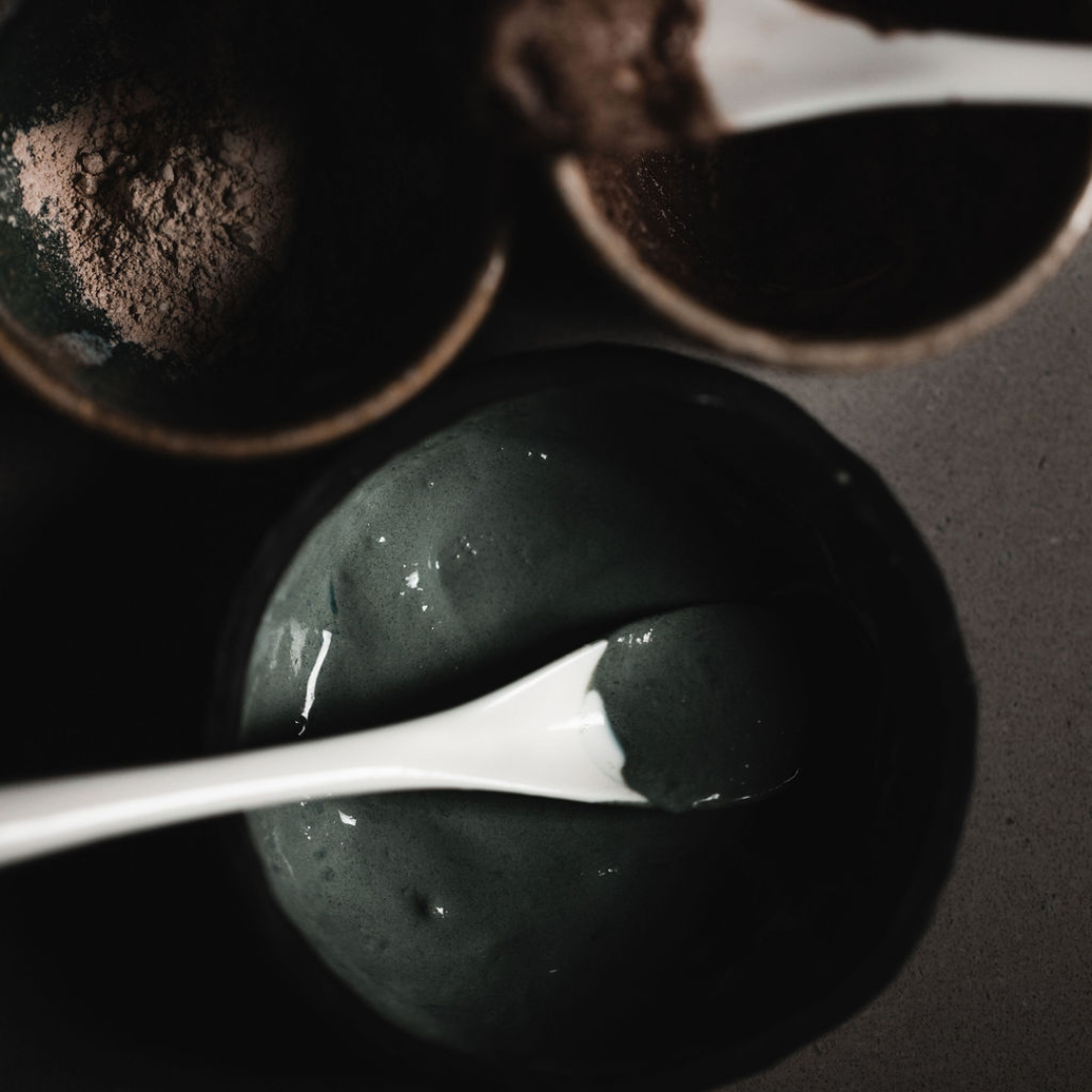 A stormy turquoise coloured paste (Oasis Black's Mineral Clay Mud Mask in Byron) in a black clay mixing bowl with a ceramic spoon, with 2 other mixing bowls. Byron is a stormy turquoise coloured mask featuring French Green Clay, Spirulina, Blue Butterfly Pea Flower, Lemon Myrtle and Black Seeds. Available exclusively from Oasis Black - makers of All Natural and Organic Botanical Skincare featuring Black Seeds (Nigella Sativa). Handmade in small batches to waterless formulations in the Byron Bay Hinterland.