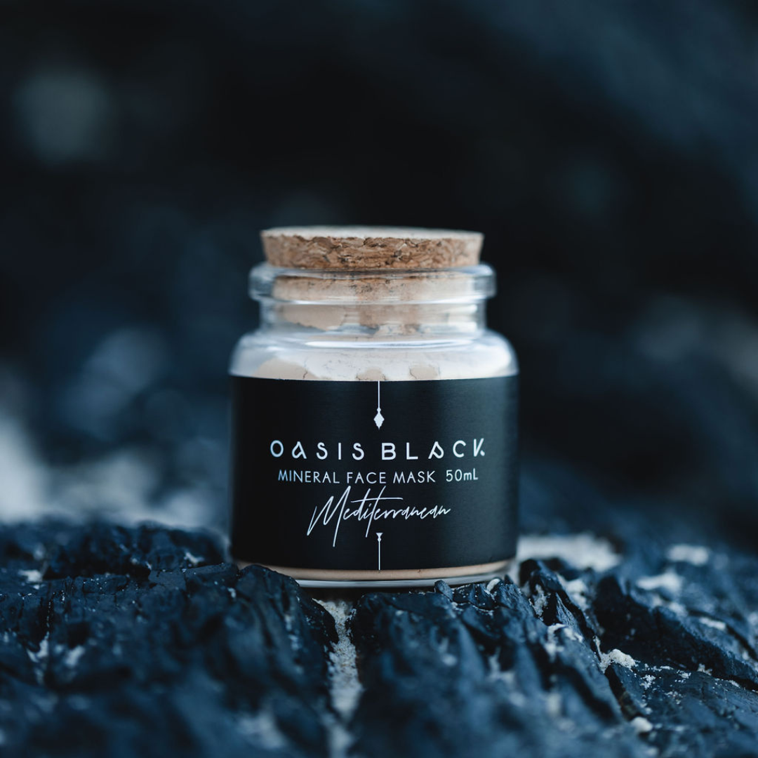 Glass jar (with a cork lid) of Oasis Black Mineral Facial Mask in Mediterranean on a dark rocky background. Mediterranean is a terracotta coloured mask activated with water and featuring Mediterranean Clays, Rose Petals, Hibiscus, Aloe Vera, Calendula and Black Seeds. Available exclusively from Oasis Black - makers of All Natural and Organic Botanical Skincare featuring Black Seeds (Nigella Sativa). Handmade in small batches to waterless formulations in the Byron Bay Hinterland. 