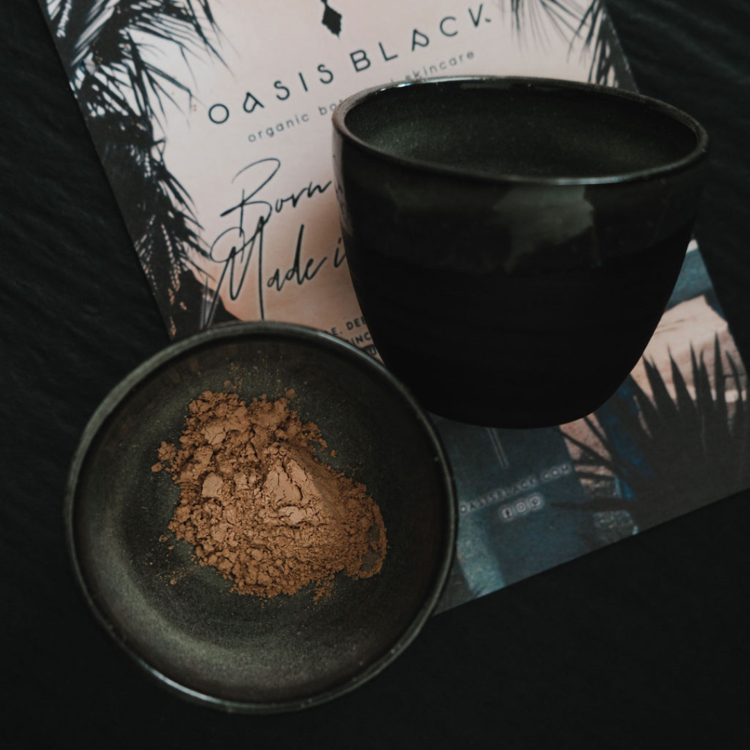 2 x Black clay cosmic mixing bowls, one with terracotta coloured powder sitting on Oasis Black branded flyer. The powder is Oasis Black's Mineral Mud Mask in Mediterranean that's activated with water & featuring Mediterranean Clays, Rose Petals, Hibiscus, Aloe Vera, Calendula & Black Seeds. Available exclusively from Oasis Black - makers of All Natural and Organic Botanical Skincare featuring Black Seeds (Nigella Sativa). Handmade in small batches to waterless formulations in the Byron Bay Hinterland. 