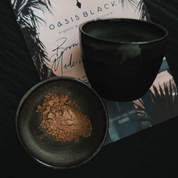 2 x Black clay cosmic mixing bowls, one with terracotta coloured powder sitting on Oasis Black branded flyer. The powder is Oasis Black's Mineral Mud Mask in Mediterranean that's activated with water & featuring Mediterranean Clays, Rose Petals, Hibiscus, Aloe Vera, Calendula & Black Seeds. Available exclusively from Oasis Black - makers of All Natural and Organic Botanical Skincare featuring Black Seeds (Nigella Sativa). Handmade in small batches to waterless formulations in the Byron Bay Hinterland. 
