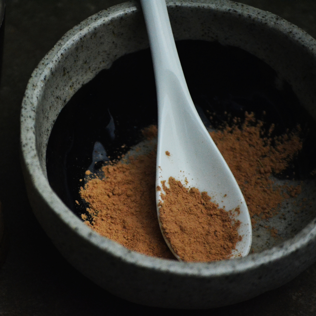 Speckled grey clay mixing bowl with terracotta coloured powder and a white ceramic teaspoon on a dark background. The powder is Oasis Black's Mineral Mud Mask in Mediterranean that's activated with water & featuring Mediterranean Clays, Rose Petals, Hibiscus, Aloe Vera, Calendula & Black Seeds. Available exclusively from Oasis Black - makers of All Natural and Organic Botanical Skincare featuring Black Seeds (Nigella Sativa). Handmade in small batches to waterless formulations in the Byron Bay Hinterland. 