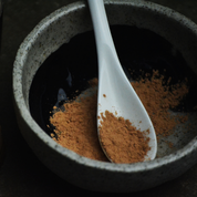 Speckled grey clay mixing bowl with terracotta coloured powder and a white ceramic teaspoon on a dark background. The powder is Oasis Black's Mineral Mud Mask in Mediterranean that's activated with water & featuring Mediterranean Clays, Rose Petals, Hibiscus, Aloe Vera, Calendula & Black Seeds. Available exclusively from Oasis Black - makers of All Natural and Organic Botanical Skincare featuring Black Seeds (Nigella Sativa). Handmade in small batches to waterless formulations in the Byron Bay Hinterland. 