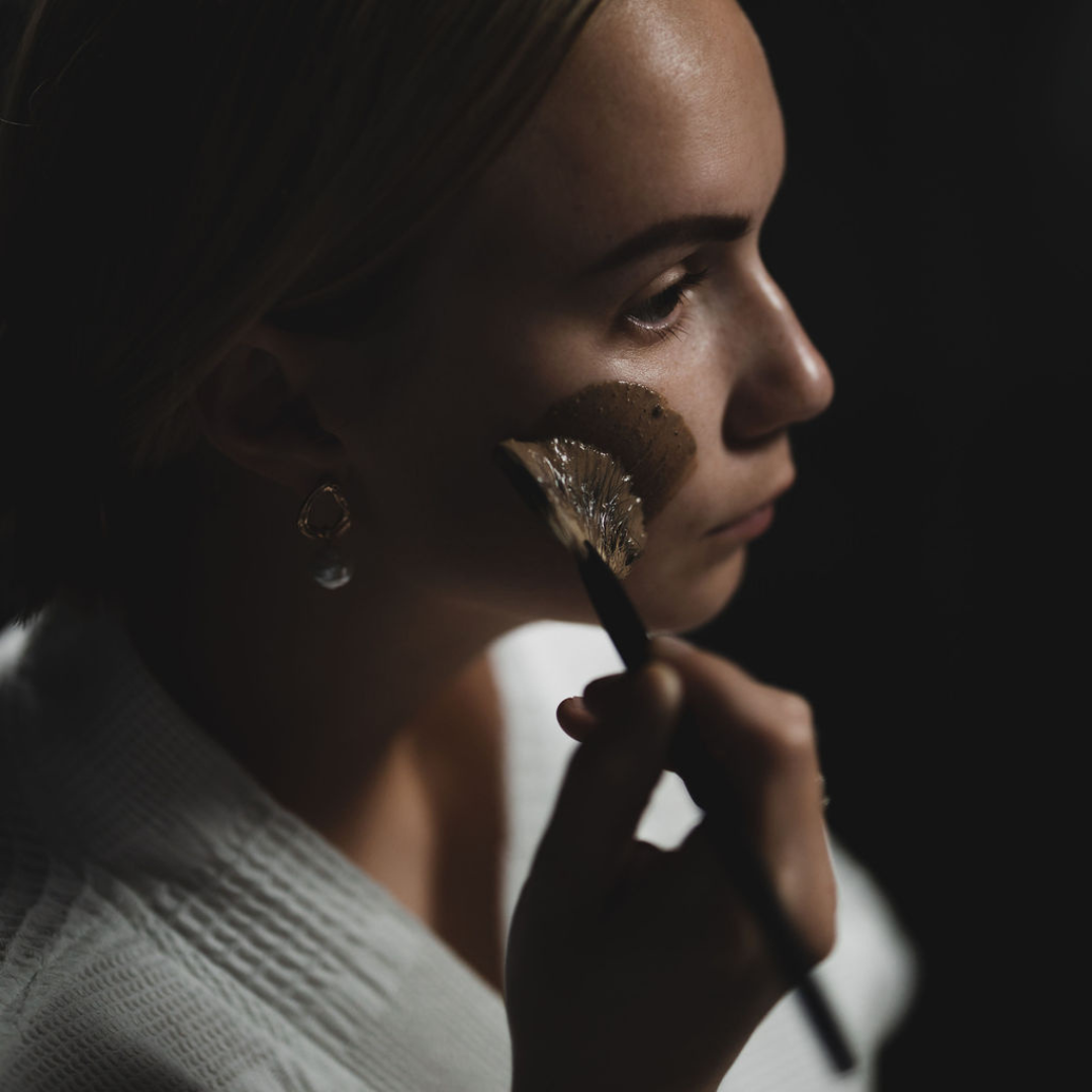 Woman applying Oasis Black's Moroccan Mineral Clay Mud Mask to her cheek with a brush on a dark background. Moroccan is a brown coloured facial mask activated with water featuring Moroccan Rhassoul Clay, Rosehips, Rose petals & Black Seeds. Available exclusively from Oasis Black - makers of All Natural and Organic Botanical Skincare featuring Black Seeds (Nigella Sativa). All products are handmade in small batches to waterless formulations in the Byron Bay hinterland