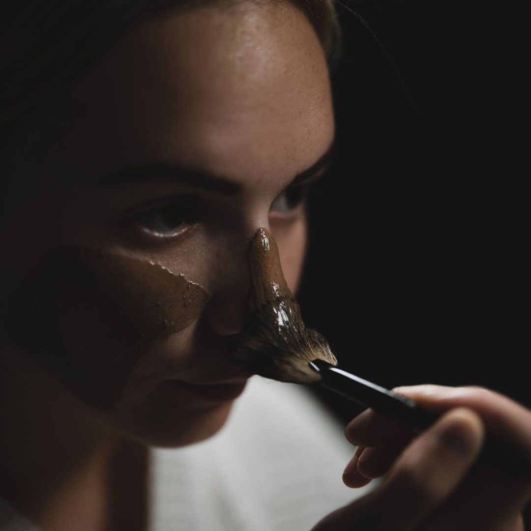 Woman applying Oasis Black's Moroccan Mineral Clay Mud Mask to her nose with a brush on a dark background. Moroccan is a brown coloured facial mask activated with water featuring Moroccan Rhassoul Clay, Rosehips, Rose petals & Black Seeds. Available exclusively from Oasis Black - makers of All Natural and Organic Botanical Skincare featuring Black Seeds (Nigella Sativa). All products are handmade in small batches to waterless formulations in the Byron Bay hinterland