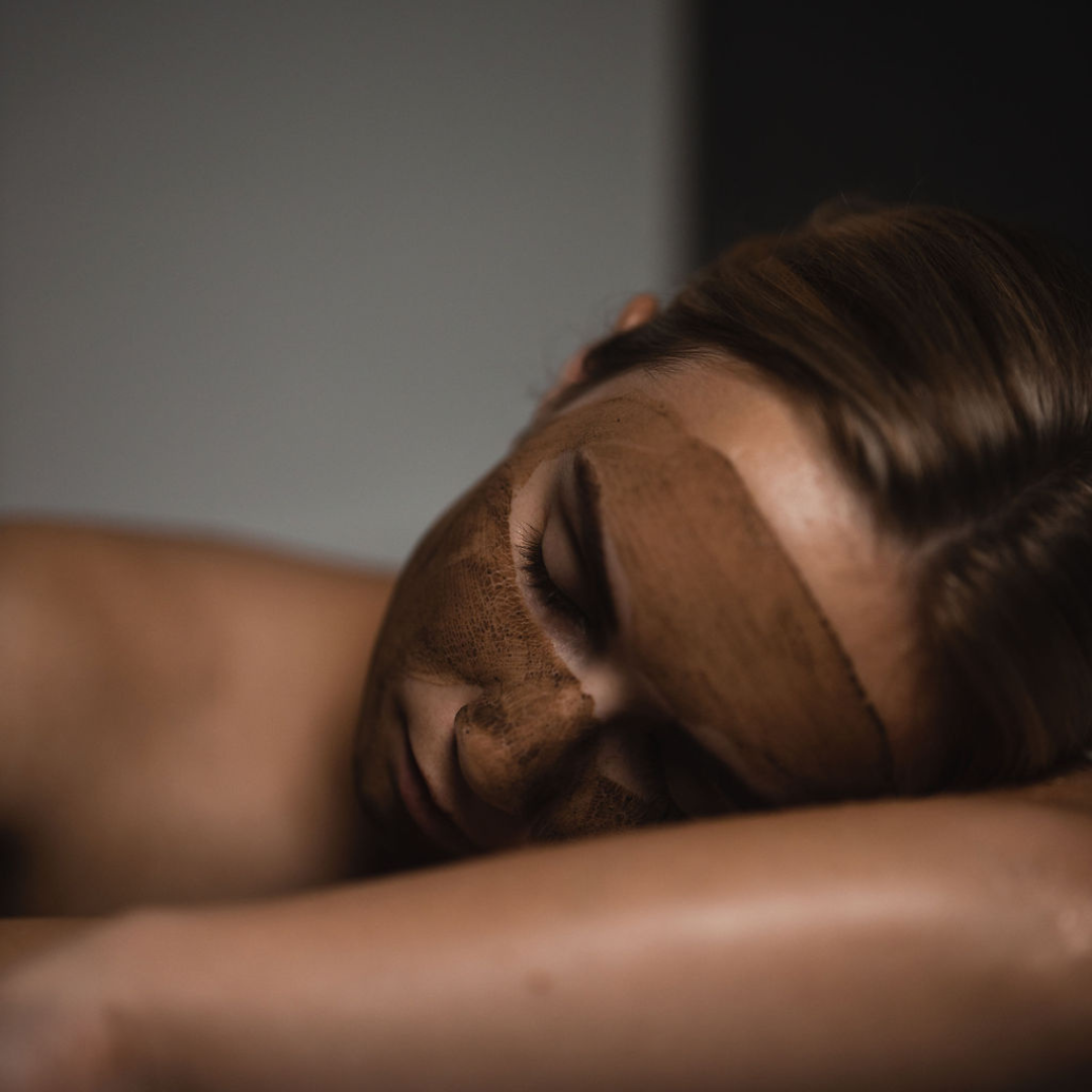 Woman with Oasis Black's Mineral Clay Mud Mask in Moroccan applied to her face, resting her head on her arm whilst it dries. Moroccan is a brown coloured facial mask activated with water featuring Moroccan Rhassoul Clay, Rosehips, Rose petals & Black Seeds. Available exclusively from Oasis Black - makers of All Natural and Organic Botanical Skincare featuring Black Seeds (Nigella Sativa). All products are handmade in small batches to waterless formulations in the Byron Bay hinterland