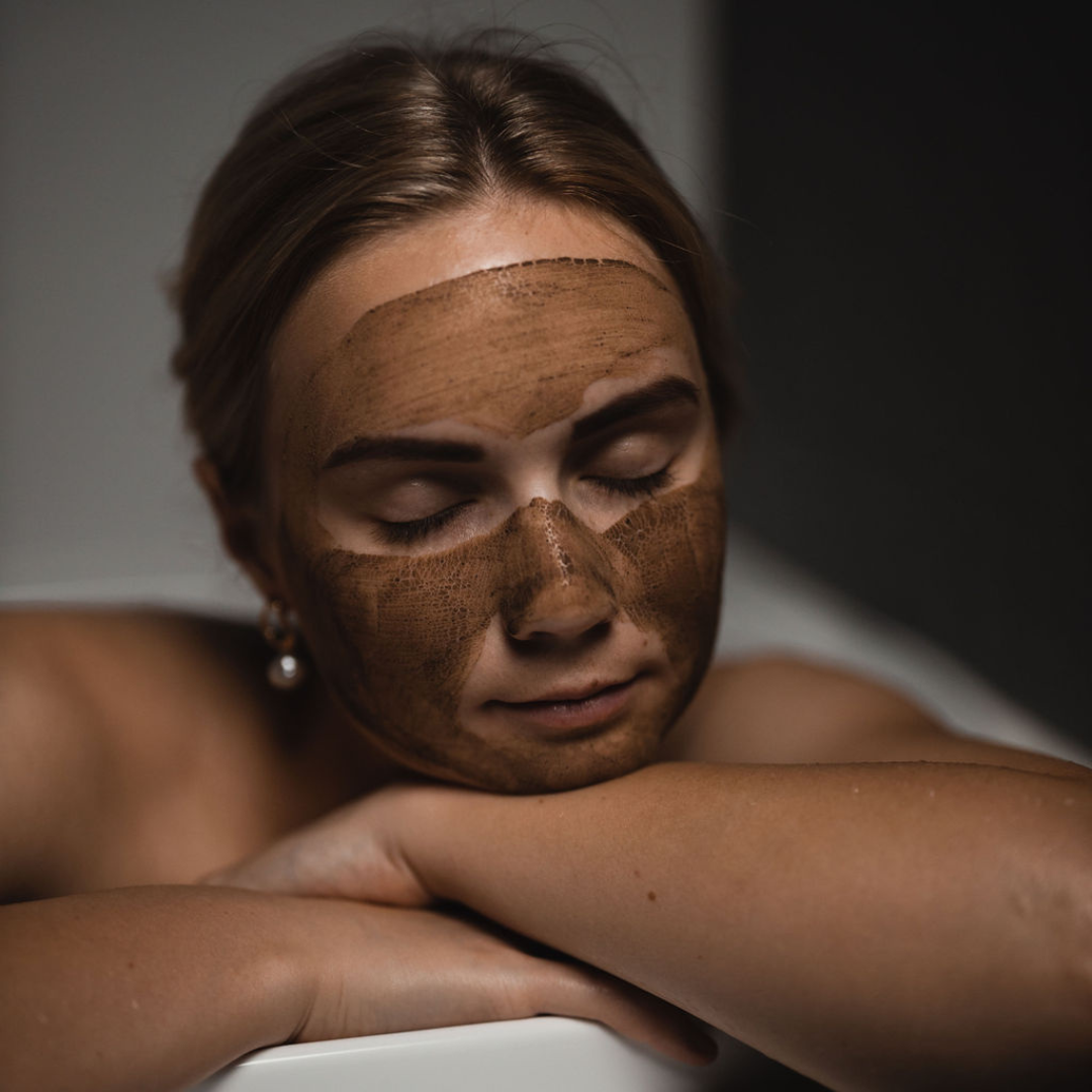 Woman with Oasis Black's Mineral Clay Mud Mask in Moroccan applied to her face, leaning and resting on the side of a bath whilst it dries. Moroccan is a brown coloured facial mask activated with water featuring Moroccan Rhassoul Clay, Rosehips, Rose petals & Black Seeds. Available exclusively from Oasis Black - makers of All Natural and Organic Botanical Skincare featuring Black Seeds (Nigella Sativa). All products are handmade in small batches to waterless formulations in the Byron Bay hinterland