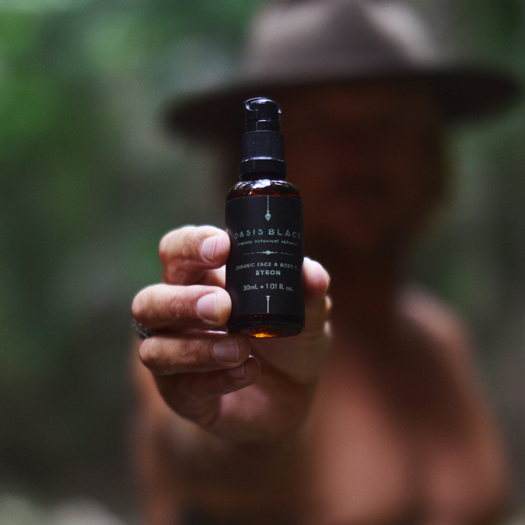 100% Organic and All Natural Face & Body Oil Featuring Black Seed Oil – BYRON – Scent inspired by Byron Bay packaged in 30ml Amber Glass Bottle pictured being held up in front of a Man wearing a hat - Non Greasy, Multi-Purpose, Fast Absorbing, Vegan Friendly, Nutrient Dense, Eco-friendly, Sustainable, Waterless and Unisex Skincare Oils made by Oasis Black – Organic Botanical Skincare - Born in Morocco, Made in small handmade batches in the Byron Bay region