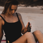 100% Organic and All Natural Face & Body Oil Featuring Black Seed Oil – BYRON – Scent inspired by Byron Bay packaged in 100ml Amber Glass Bottle with Natural Golden Mica pictured being held on the knee of a woman sitting on the beach - Non Greasy, Multi-Purpose, Fast Absorbing, Vegan Friendly, Nutrient Dense, Eco-friendly, Sustainable, Waterless and Unisex Skincare Oils made by Oasis Black – Organic Botanical Skincare - Born in Morocco, Made in small handmade batches in the Byron Bay region