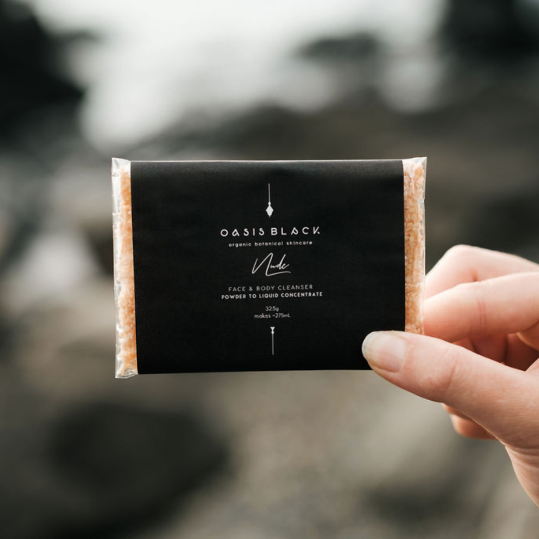 Hand holding a packet of Oasis Black's Organic & All Natural Face & Body Cleanser Featuring Black Seed Oil – Full Size Sachet pictured in Nude. This is a Powder to Liquid Castile Soap Cleanser Concentrate and Multipurpose Cleanser suitable for your Face, Hands, Feet and Body. The packaging is Zero Waste, Plastic Free, and Home Compostable. Oasis Black Organic Botanical Skincare products are Vegan Friendly, Waterless, Unisex, Travel Friendly.