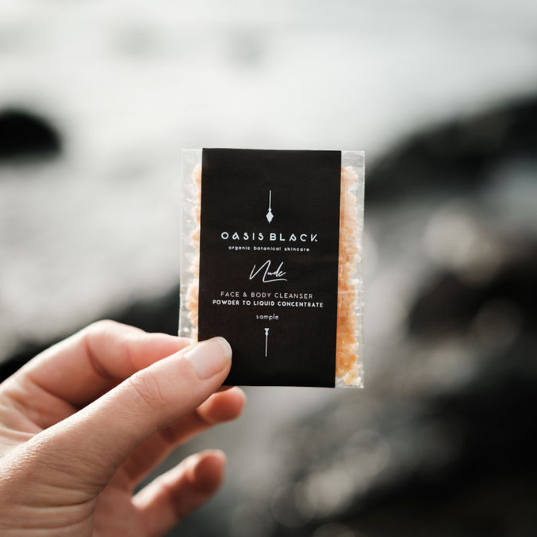 Hand holding a small packet of Oasis Black's Organic & All Natural Face & Body Cleanser Featuring Black Seed Oil – Sample Size Sachet pictured in Nude. This is a Powder to Liquid Castile Soap Cleanser Concentrate and Multipurpose Cleanser suitable for your Face, Hands, Feet and Body. The packaging is Zero Waste, Plastic Free, and Home Compostable. Oasis Black Organic Botanical Skincare products are Vegan Friendly, Waterless, Unisex, Travel Friendly.