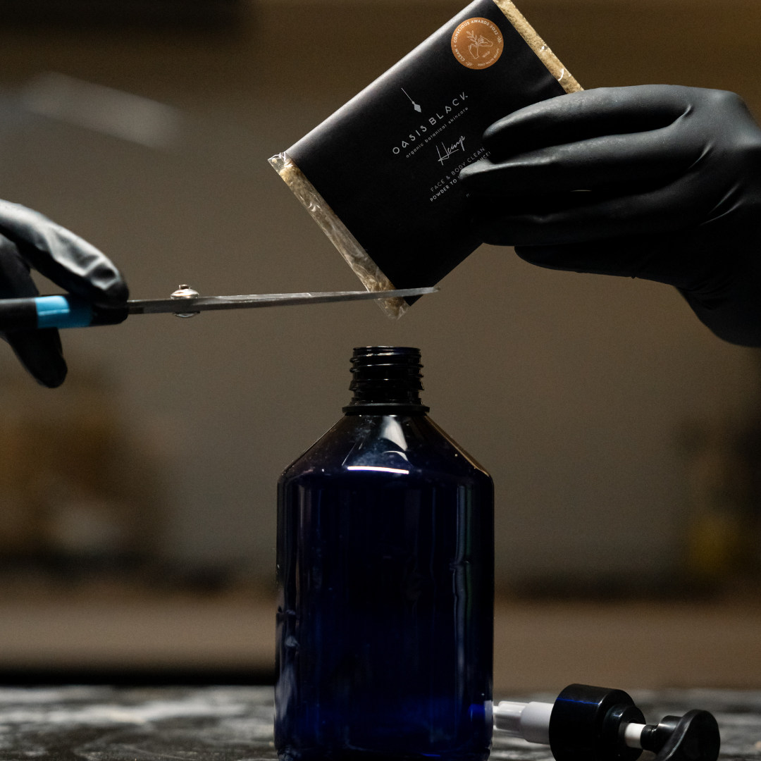 Person cutting a package of Oasis Black's Face & Body, Powder to Liquid Cleanser Concentrate in HEMP with scissors over a blue bottle. Oasis Black's Cleanser Concentrates are All natural, zero waste castile soap based powdered cleansers that turn into liquid face and body wash with water. Available exclusively from Oasis Black - makers of All Natural and Organic Botanical Skincare featuring Black Seeds (Nigella Sativa). Handmade in small batches to waterless formulations in the Byron Bay Hinterland.