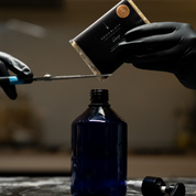 Person cutting a package of Oasis Black's Face & Body, Powder to Liquid Cleanser Concentrate in HEMP with scissors over a blue bottle. Oasis Black's Cleanser Concentrates are All natural, zero waste castile soap based powdered cleansers that turn into liquid face and body wash with water. Available exclusively from Oasis Black - makers of All Natural and Organic Botanical Skincare featuring Black Seeds (Nigella Sativa). Handmade in small batches to waterless formulations in the Byron Bay Hinterland.