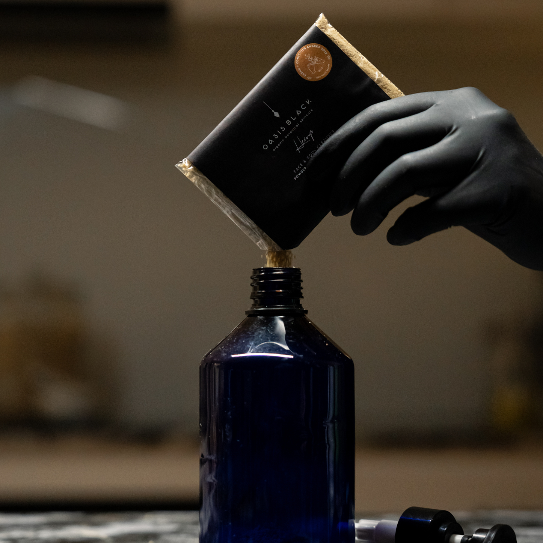Person pouring a package of Oasis Black's Face & Body, Powder to Liquid Cleanser Concentrate in HEMP into a blue bottle. Oasis Black's Cleanser Concentrates are All natural, zero waste castile soap based powdered cleansers that turn into liquid face and body wash with water. Available exclusively from Oasis Black - makers of All Natural and Organic Botanical Skincare featuring Black Seeds (Nigella Sativa). Handmade in small batches to waterless formulations in the Byron Bay Hinterland.